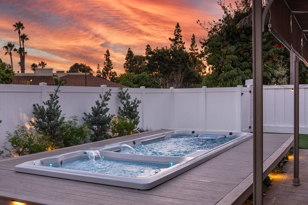 A swim spa in a backyard against a white fence and pink sunset background