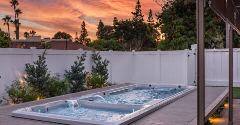 A swim spa in a backyard against a white fence and pink sunset background