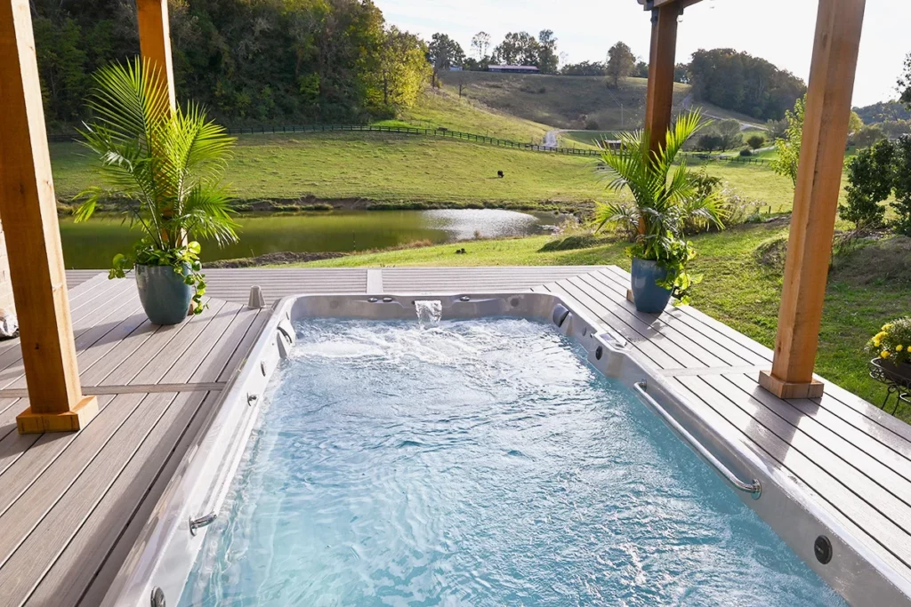 An outdoor swim spa in a deck, surrounded by green grass and shaded with a pergola