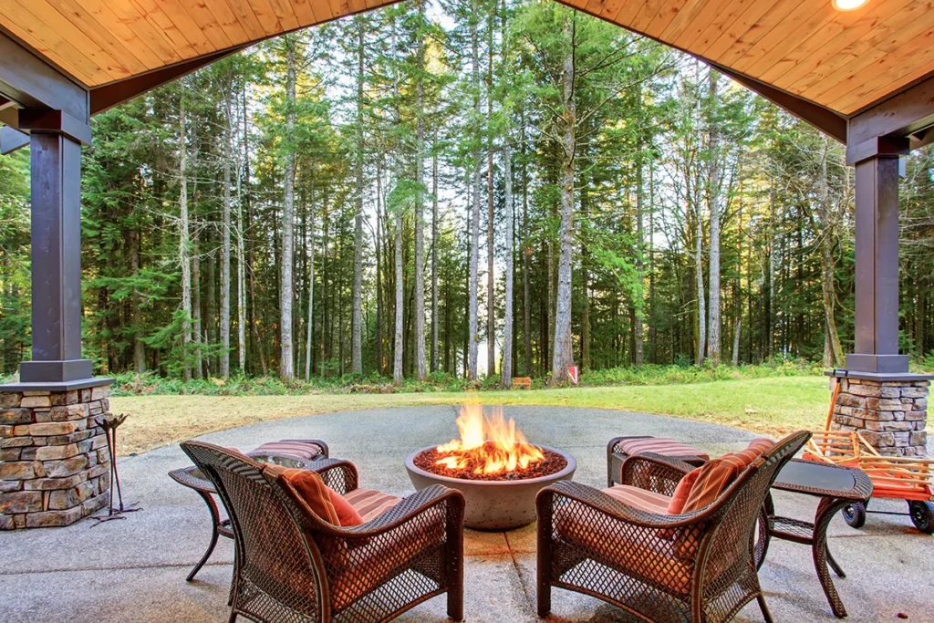 An outdoor patio in a backyard, with a stone firepit, surrounded by grass and trees