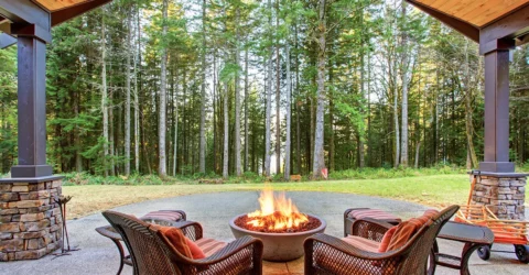 An outdoor patio in a backyard, with a stone firepit, surrounded by grass and trees