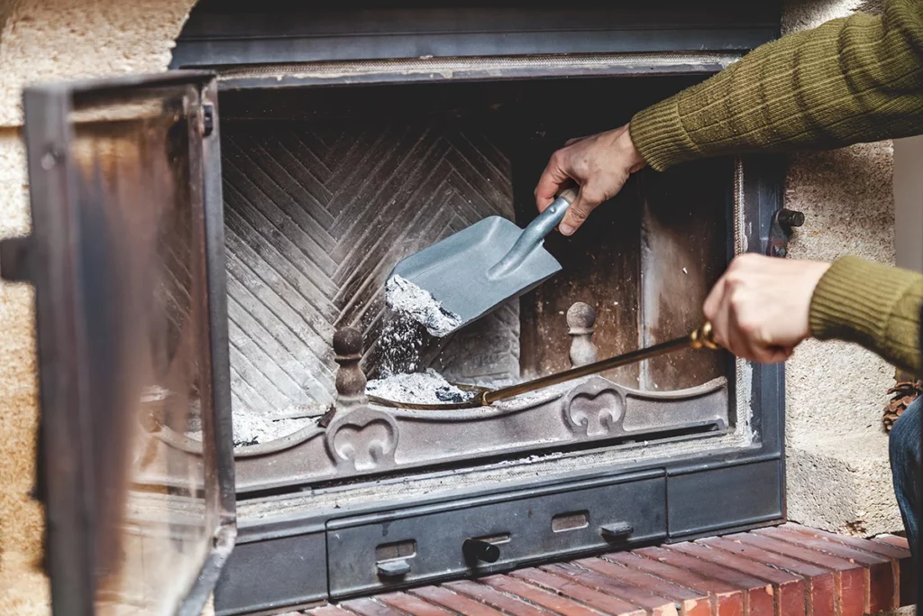 A close up of an arm in a green sweater, properly removing the ashes from a fireplace using an ash shovel