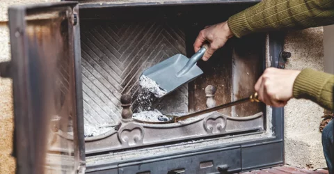 A close up of an arm in a green sweater, properly removing the ashes from a fireplace using an ash shovel