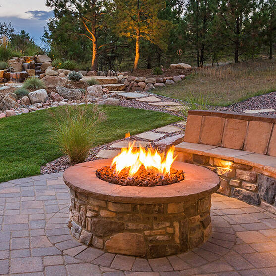 A stone firepit in a backyard in the evening, surrounded by trees and green grass.