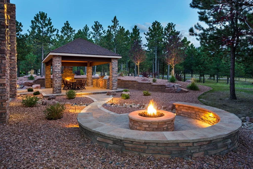 A backyard patio and outdoor dining area at night, with an outdoor fireplace and a stone firepit, with trees in the background