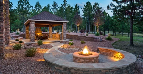 A backyard patio and outdoor dining area at night, with an outdoor fireplace and a stone firepit, with trees in the background