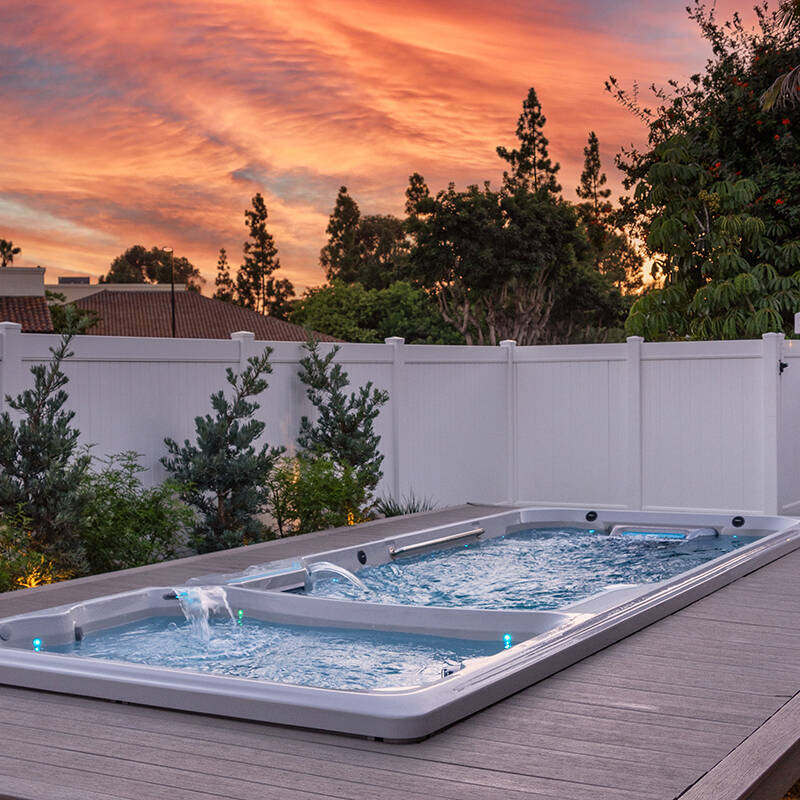 A swim spa in a backyard against a white fence and pink sunset background