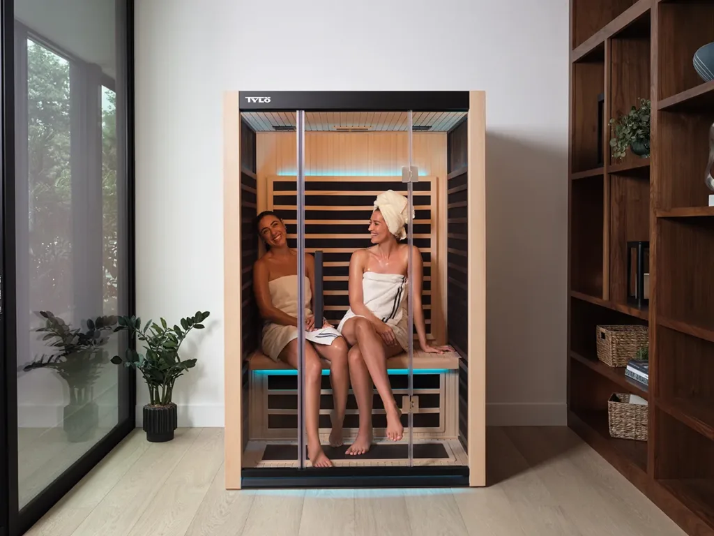 Two women in towels sit inside an infrared sauna, smiling and talking